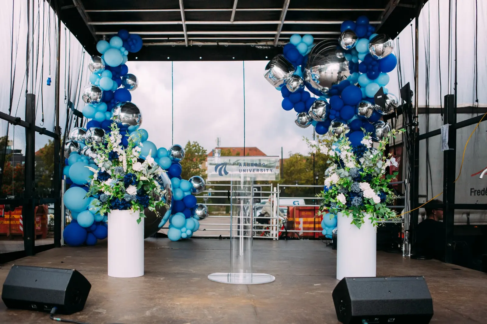 Udendørs scene til studiestart på AAU København med blå ballondekorationer, spejlkugler, blomster på hvide søjler og talerpult i akryl