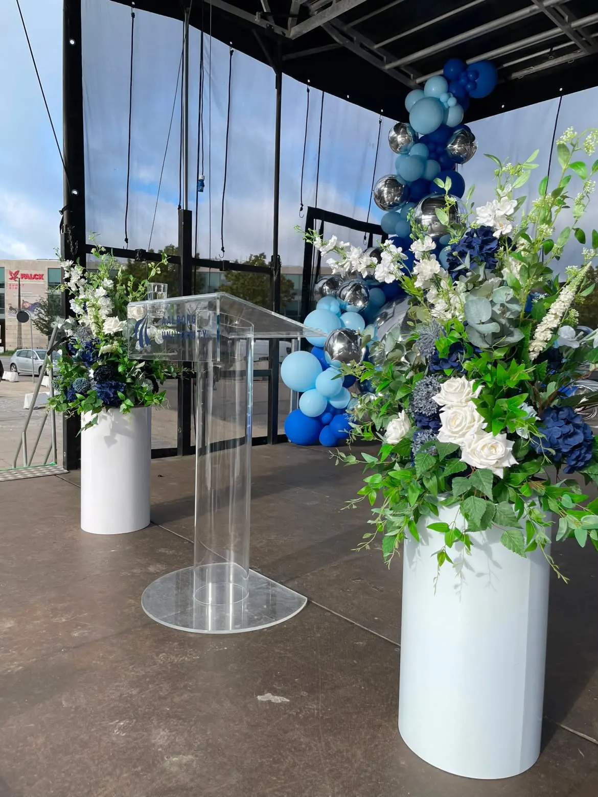 Talerpult i akryl med Aalborg University-logo mellem to blomsteropsætninger på scenen