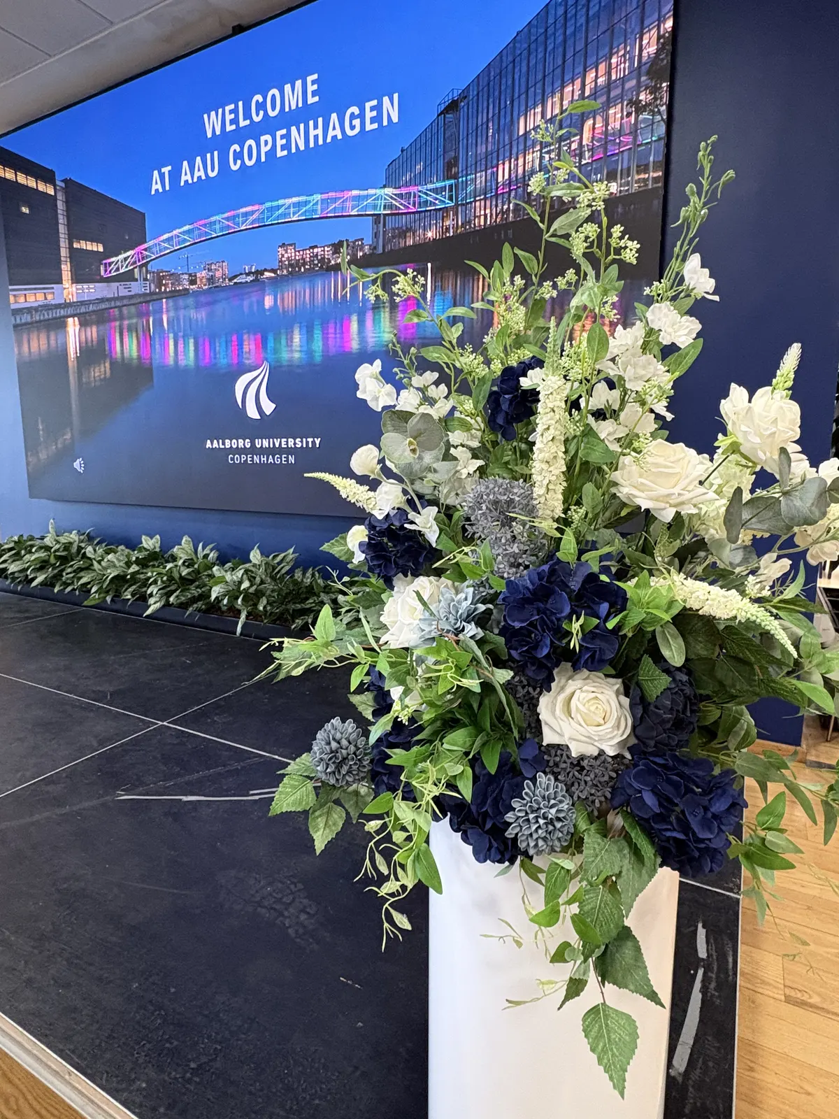 Blomsteropsætning i hvid, blå og grøn foran velkomstskærm med teksten Welcome at AAU Copenhagen