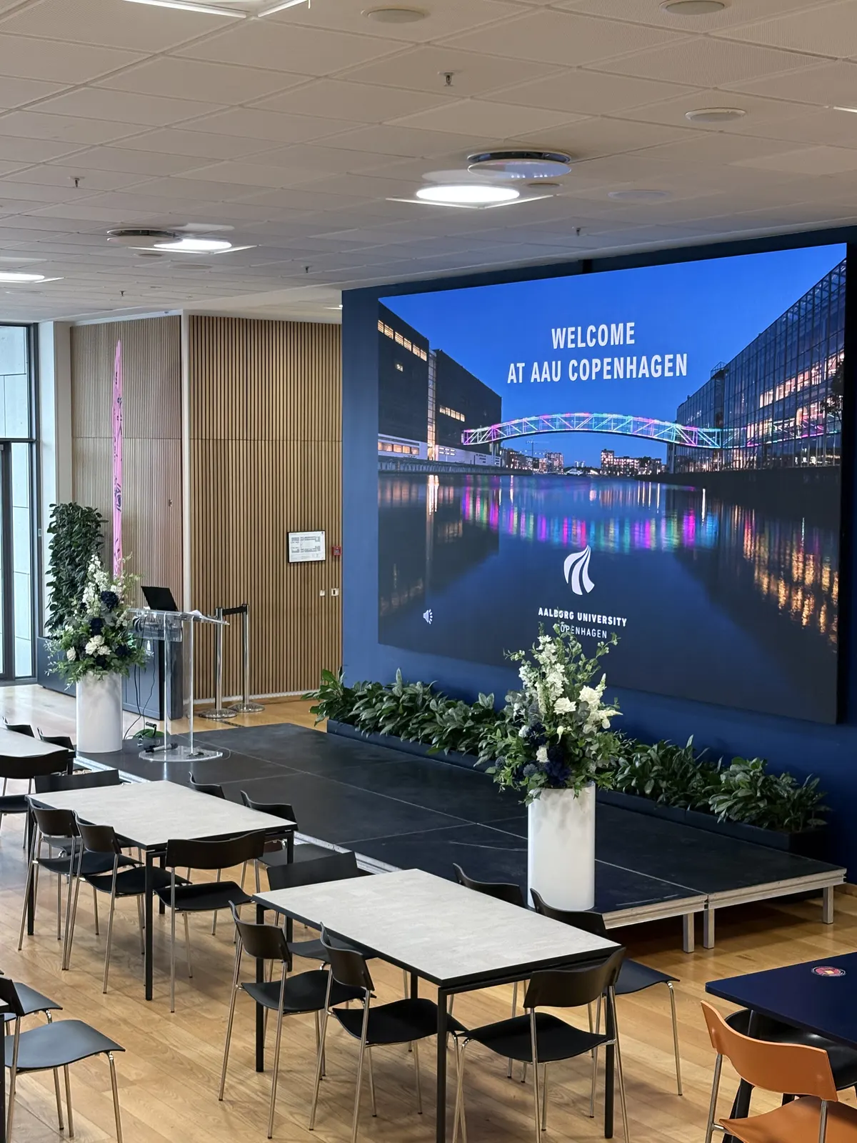 Indendørs scene på AAU København med blomsteropsætninger på hvide søjler og stor velkomstskærm