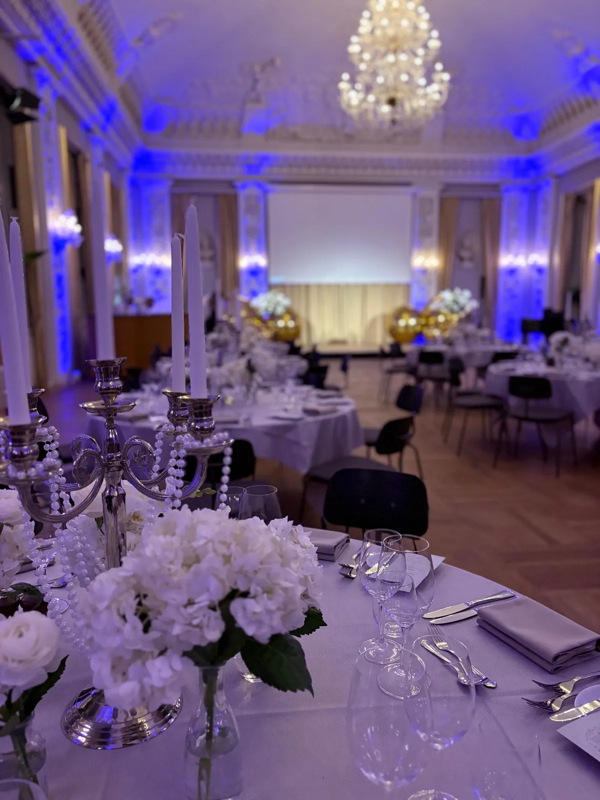 Festsalen i Moltkes Palæ med lysekrone, blå lyssætning og borde med sølvlysestager og hvide blomster