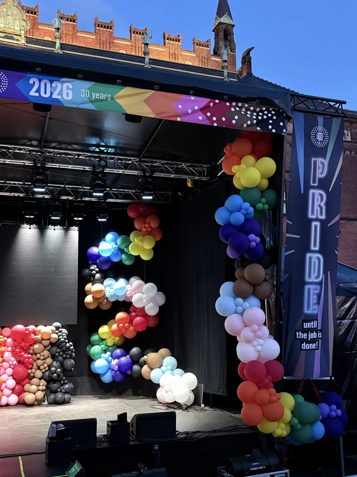 Scenen i dagslys med ballonguirlande ved Pride-banneret og ballonvæg under skærmen