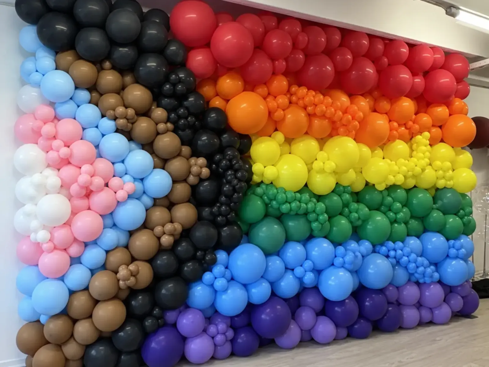 Ballonvæg i Progress Pride-farverne med regnbuestriber og lyseblå, lyserøde, hvide, brune og sorte balloner